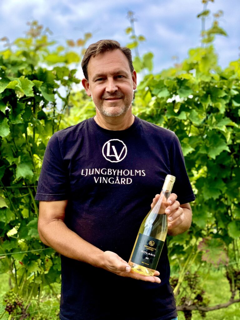 Man holding a bottle of white wine with vines in the background.