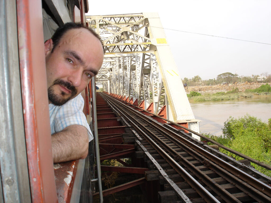 Man with head outside train window as train passes a bridge over a river.