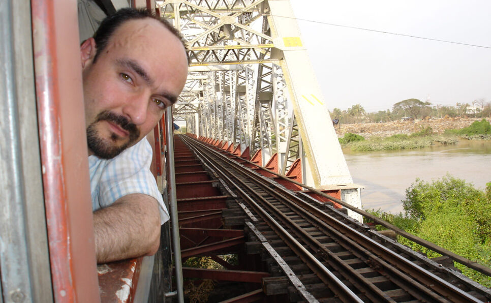 Man with head outside train window as train passes a bridge over a river.