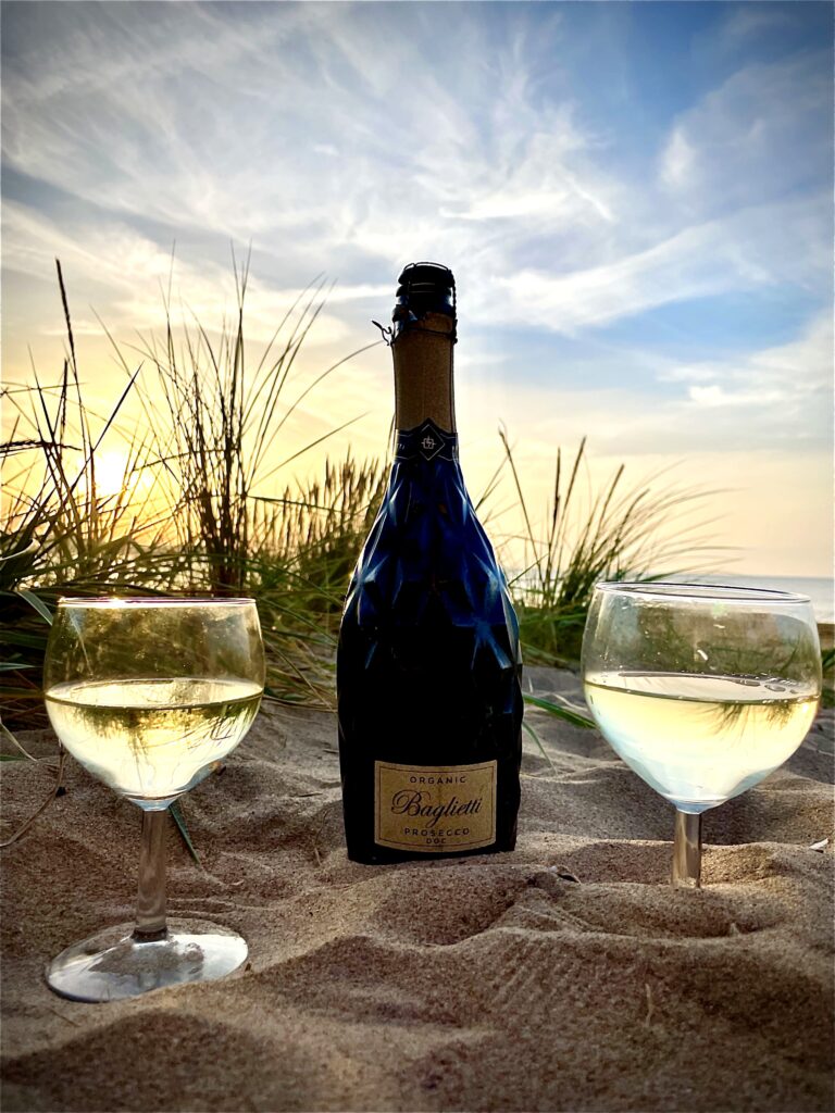 A bottle of organic Prosecco with two glasses on a sunset beach.
