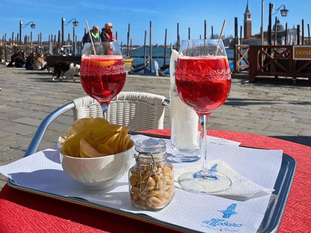 Two glasses of red coloured aperatives on a table near water