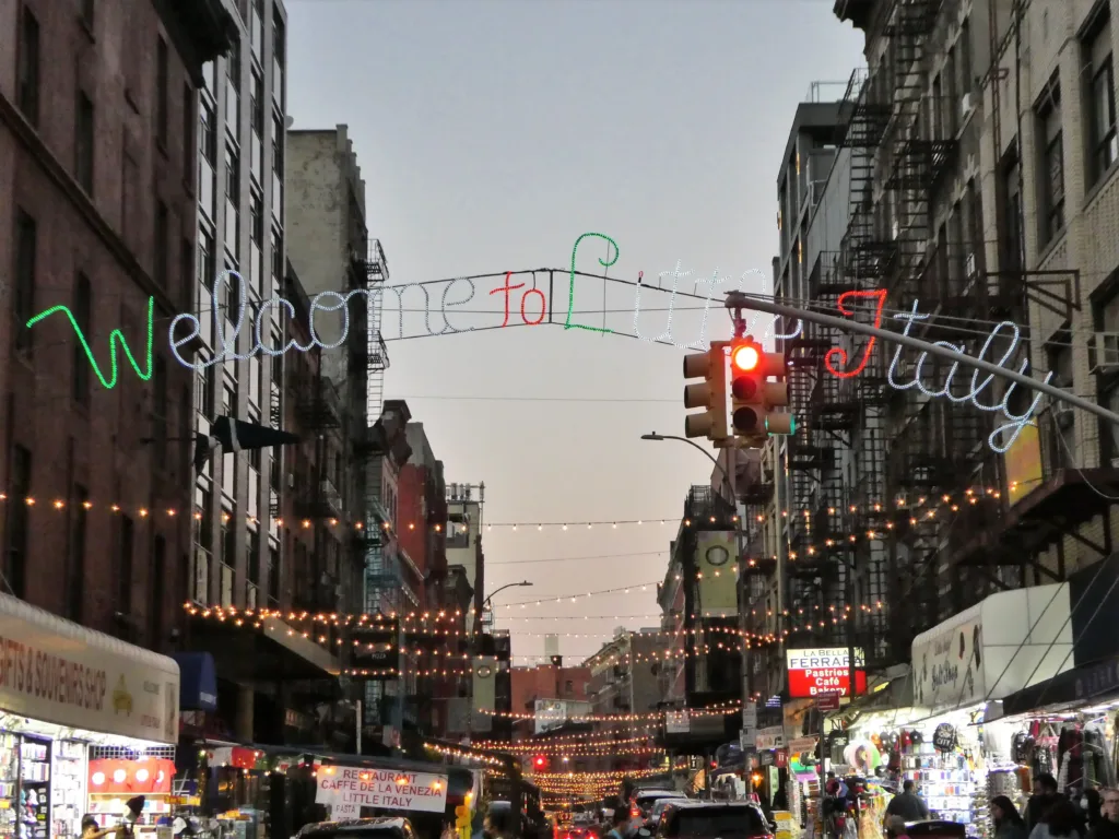 A red and green neon sign stating Welcome to Little Italy, hanging over a  lively street.