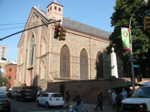 A brick church bathing in sunlight, with people and cars in front.