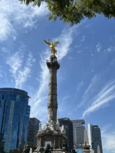 Monument with gilded statue on top amid a backdrop of high rise buildings. 