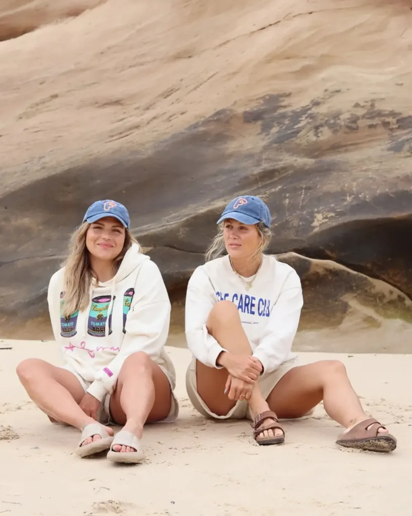 Two women sitting on the ground in white sweaters and sandals, with a big rock in the background.