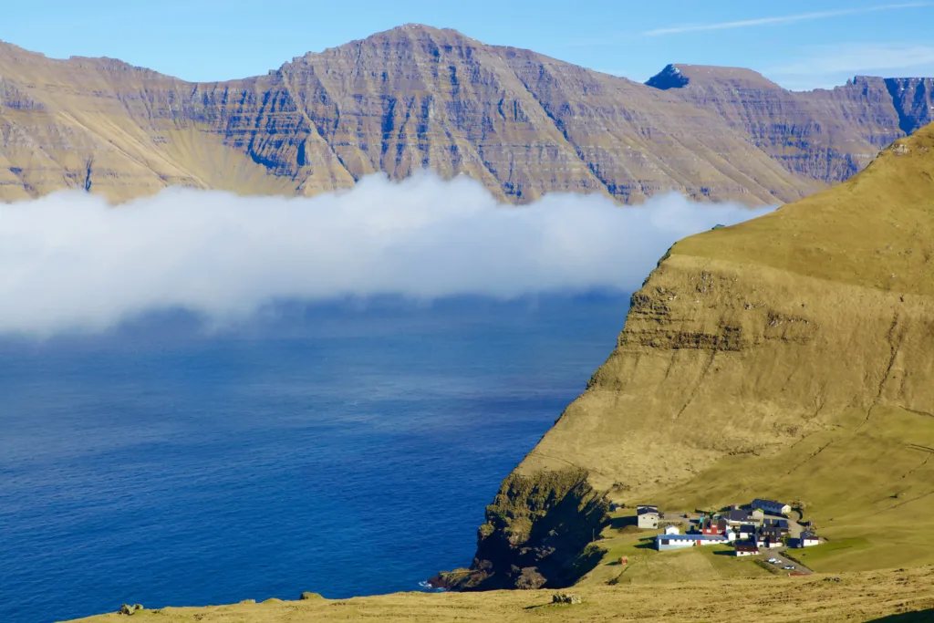 Collection of houses near a cliff with green mountains and the sea in the background.