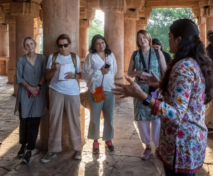 Group of women standing in a building with pillars, listening to another woman talk. 