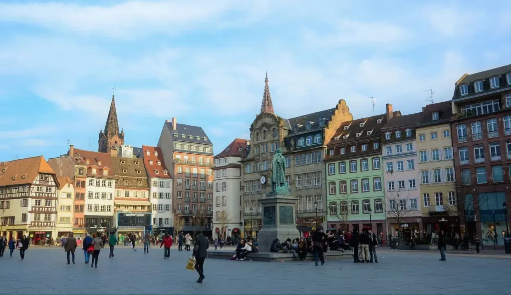 Square with statue in the middle, people sitting and walking and colourful multistory houses framing the square.