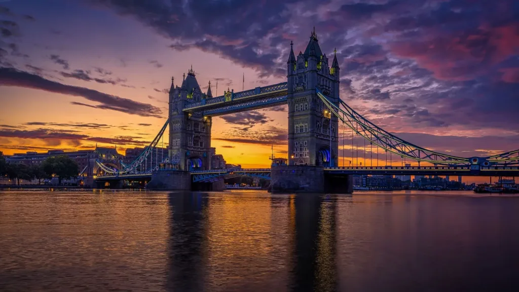 Suspension bridge with two tower connected by a walkway, spanning a river at sunset.