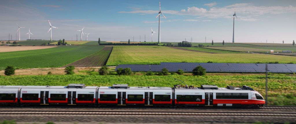 Red and white train riding past green fields with solar and wind energy installations.