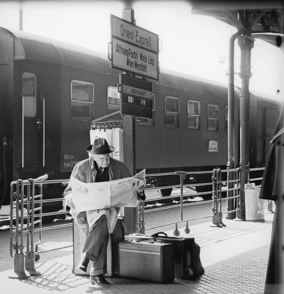 Black and white photo of older man reading the newspaper at train platform.