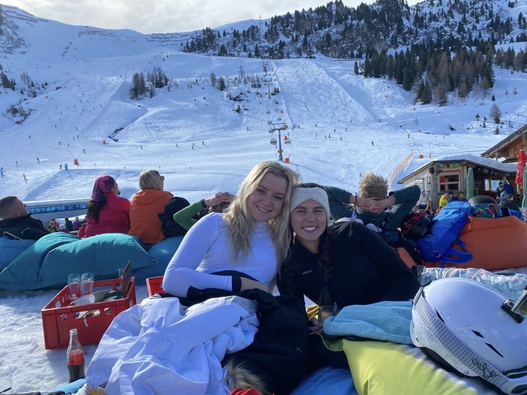 Two women sitting at the base of a ski slope enjoying refreshments