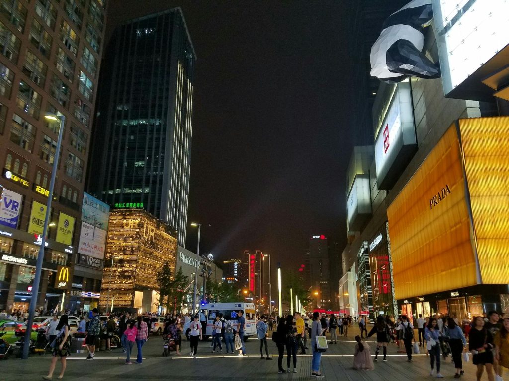 People walking no pedestrian street lined by shops and tall buildings at night