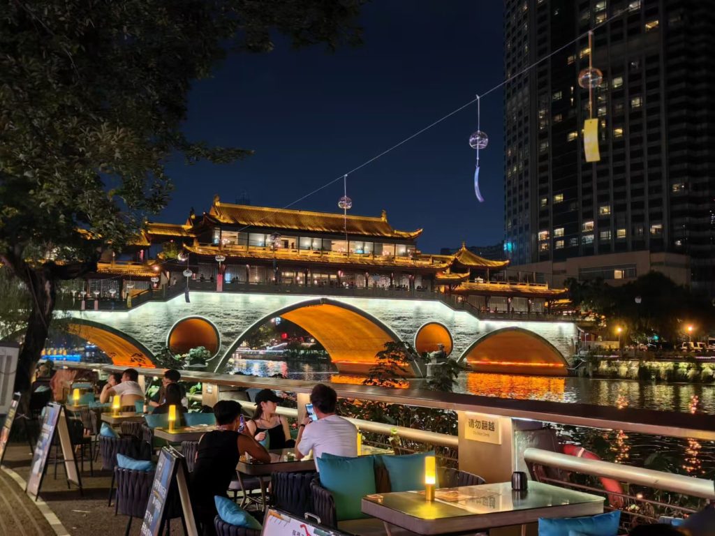 A covered bridge lit up during the evening with people sitting at tables near the river