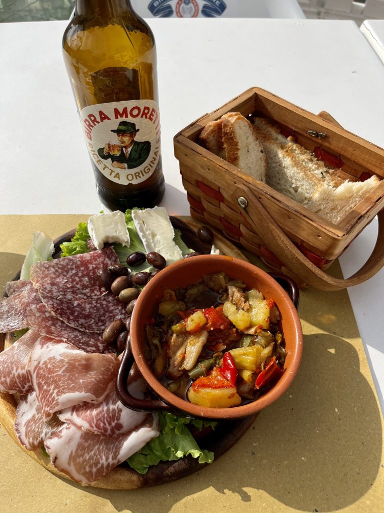 Plate of cold cuts, olives, vegetables, and bread next to a bottle of beer