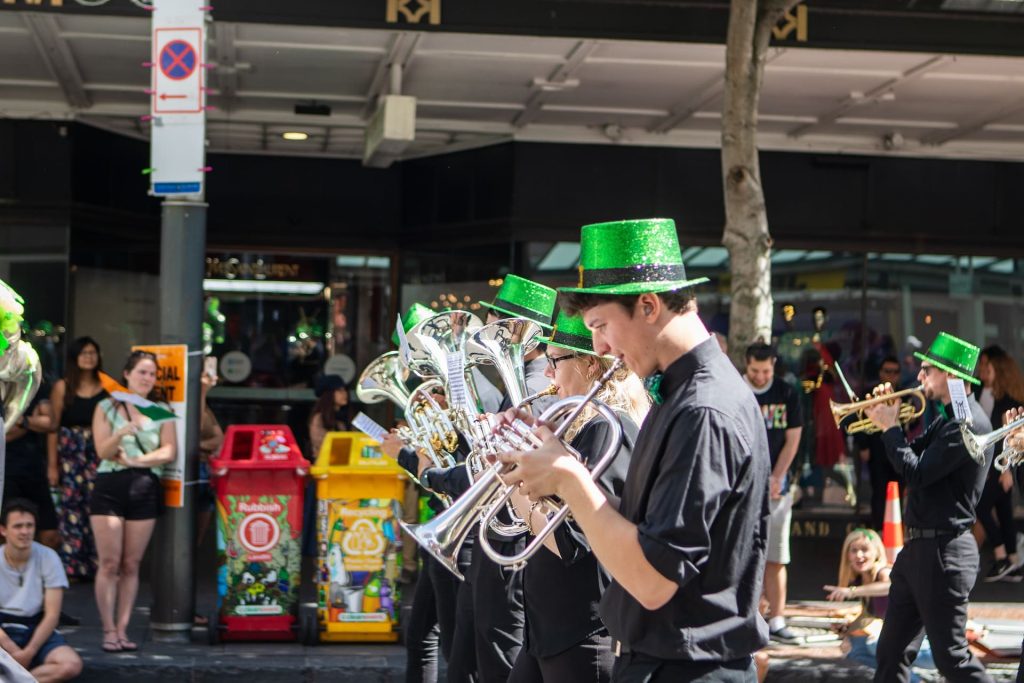 Part of a parade wearing green hats playing trumpets while an audience looks on from the street