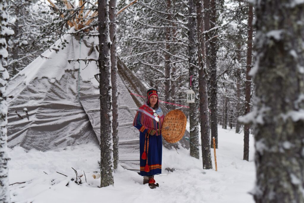 Woman with wooden drum in indigenous dress posing outside tent in the snow