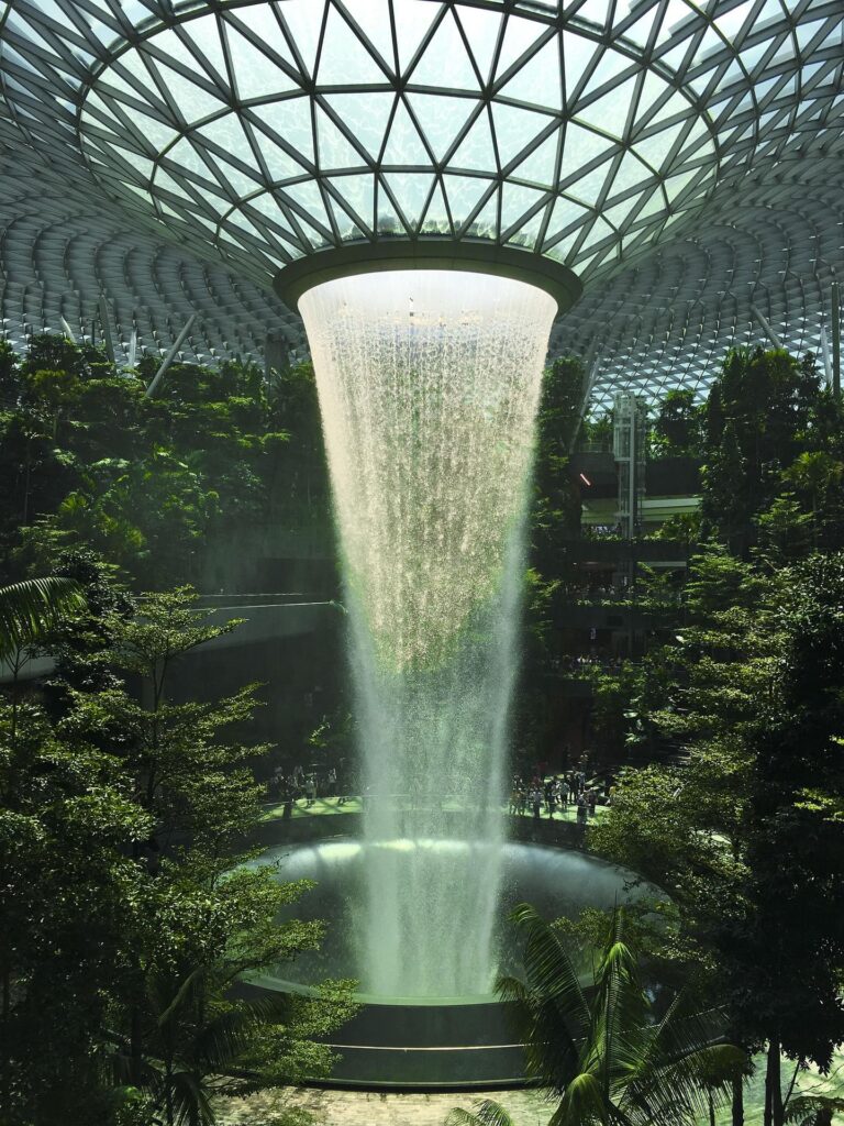 A waterfall coming out of a hole in a meshed ceiling surrounded by lush vegetation.
