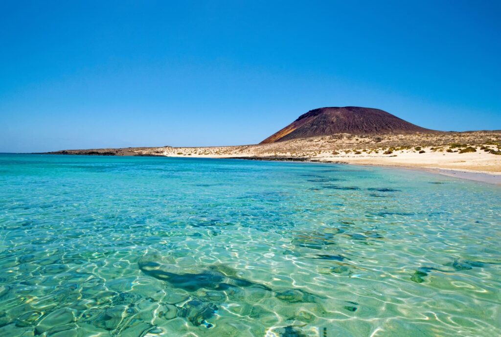 Crystal clear blue water and a beach with a hill.