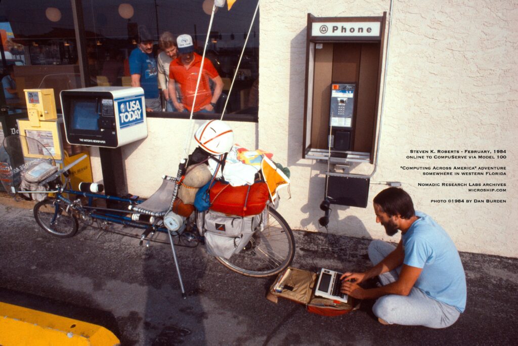 Man sitting on the ground near a payphone, working on a computer while people watch from the building window