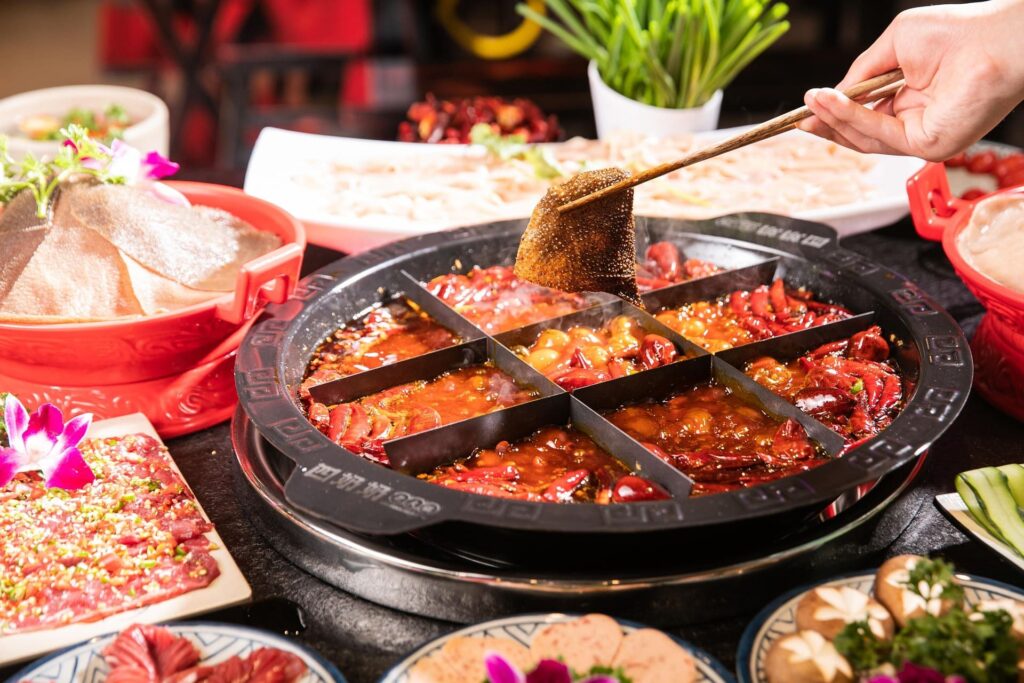 hand with chopsticks dipping food into a hotpot with side dishes surrounding it.