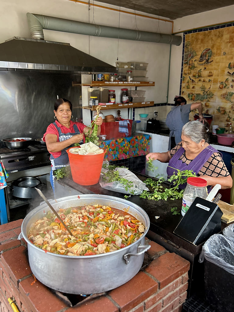 Women preparing and cooking food in a kitchen with large pots on an open fire